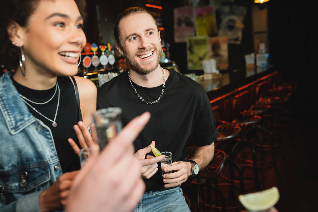 Smiling bearded man holding tequila shot near multiethnic friends in bar  - Photo, Image