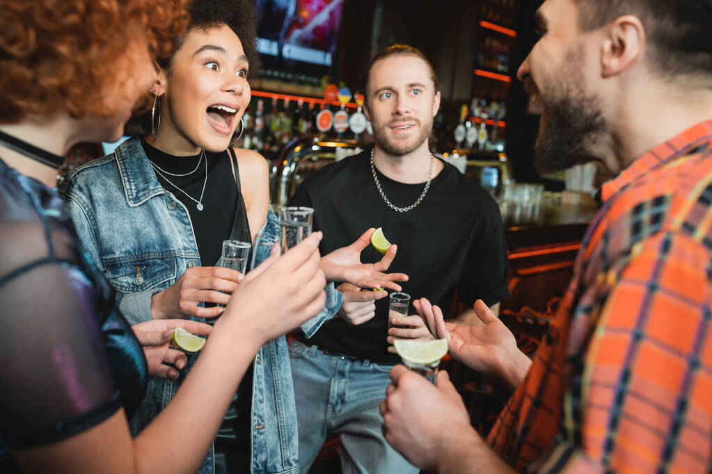 Excited african american woman holding tequila shot near blurred friends in bar  - Photo, Image