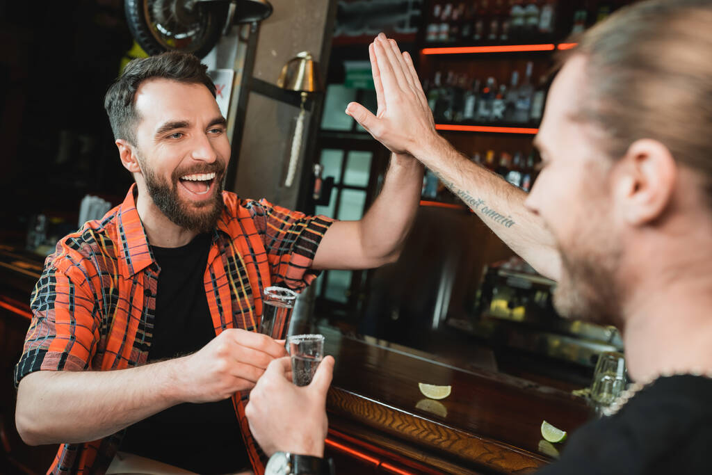 Allegro uomini barbuti dando il cinque e tenendo bicchieri di tequila nel bar  - Foto, immagini
