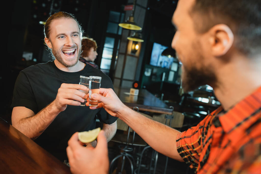 glimlachende man met baard klinkende tequila shots met wazig vriend in bar  - Foto, afbeelding