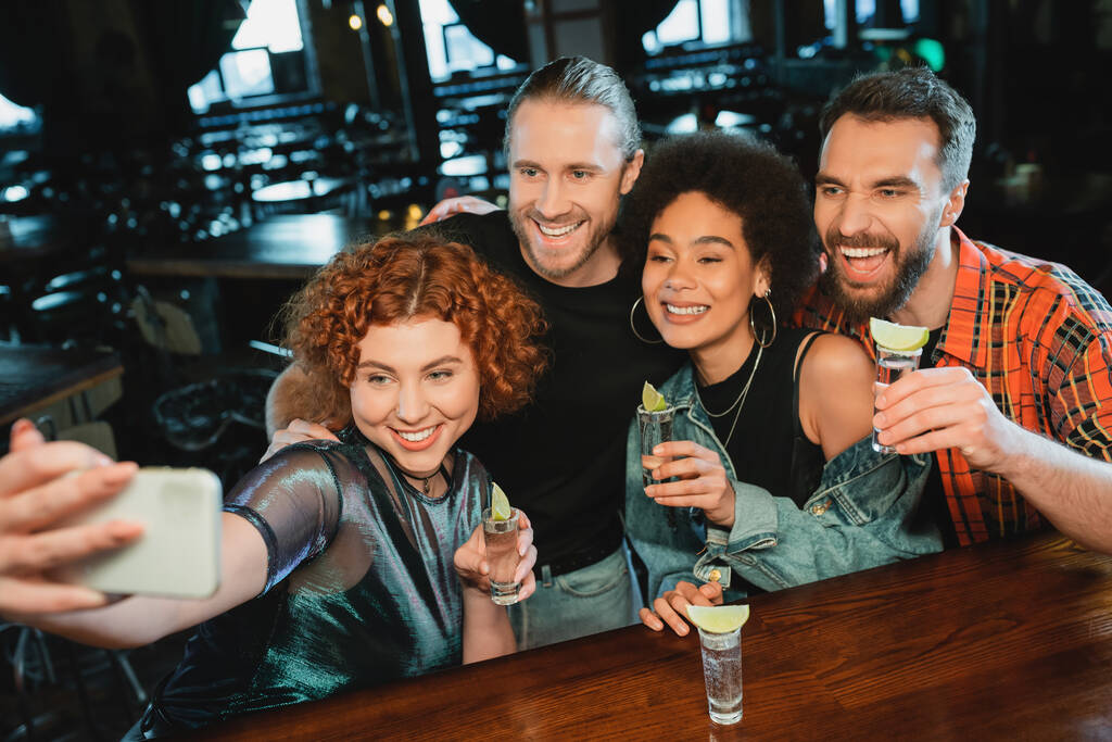 Χαρούμενοι διαφυλετικοί φίλοι με σφηνάκια τεκίλας που βγάζουν selfie στο smartphone στο μπαρ  - Φωτογραφία, εικόνα