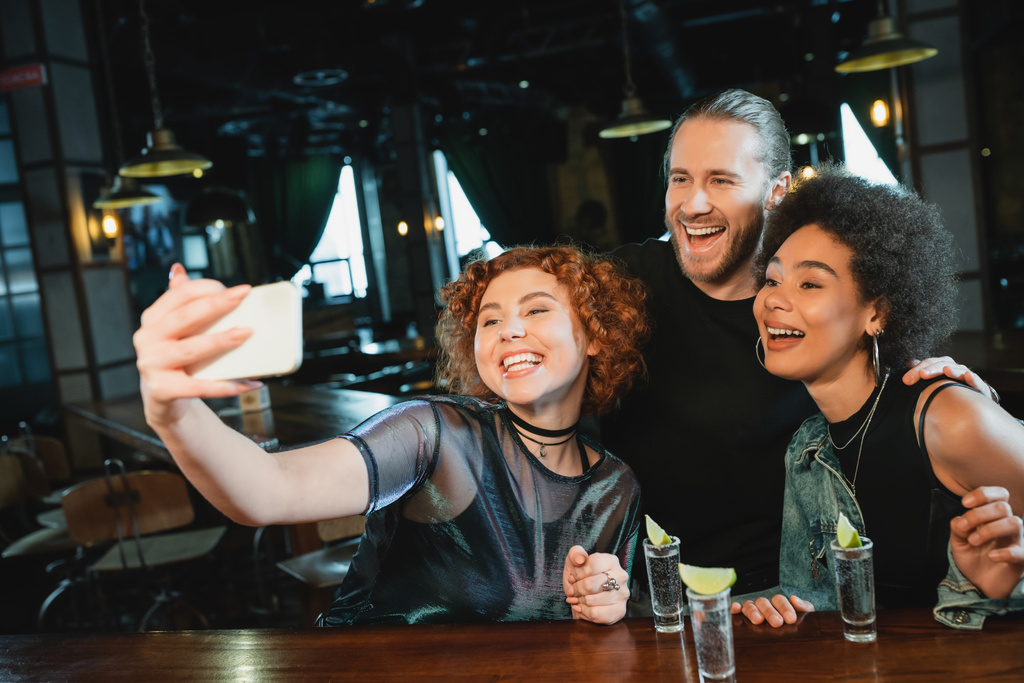 Рыжая женщина делает селфи с улыбающимися друзьями возле текилы в баре - Фото, изображение