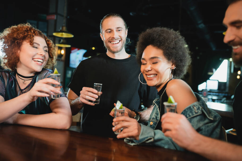 Cheerful multiethnic friends holding tequila shots with lime in bar  - Photo, Image