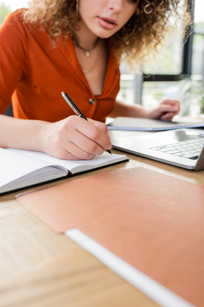 abgeschnittene Ansicht einer lockigen Geschäftsfrau, die Notizen in einem Notizbuch neben dem Laptop auf dem Schreibtisch macht  - Foto, Bild