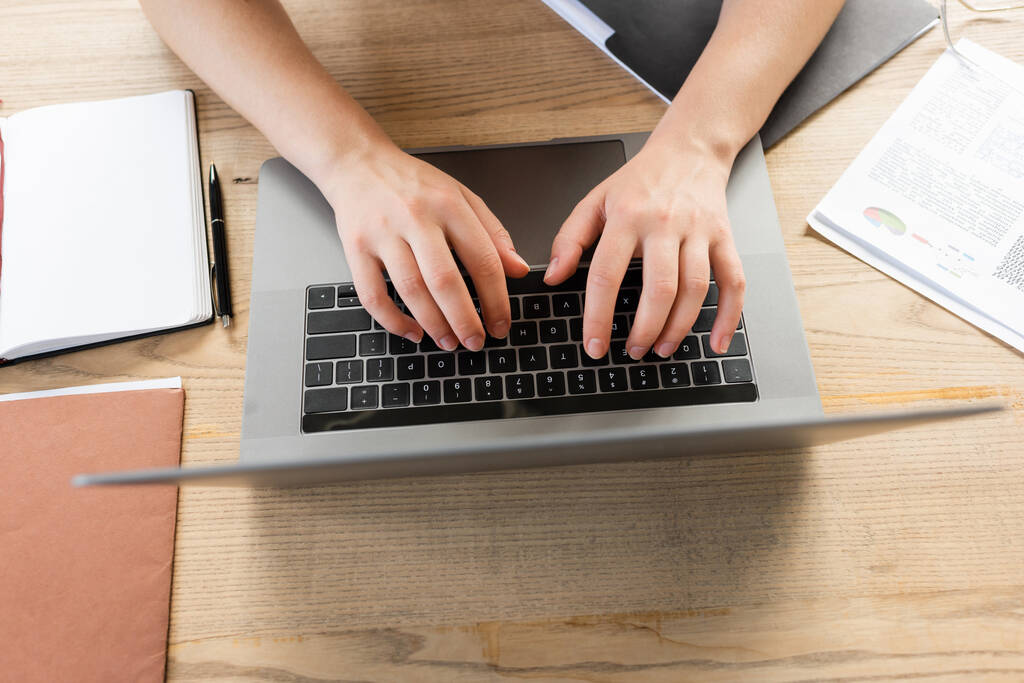 vista superior de la mujer recortada escribiendo en el teclado del ordenador portátil cerca de carpetas e infografías en el escritorio  - Foto, imagen