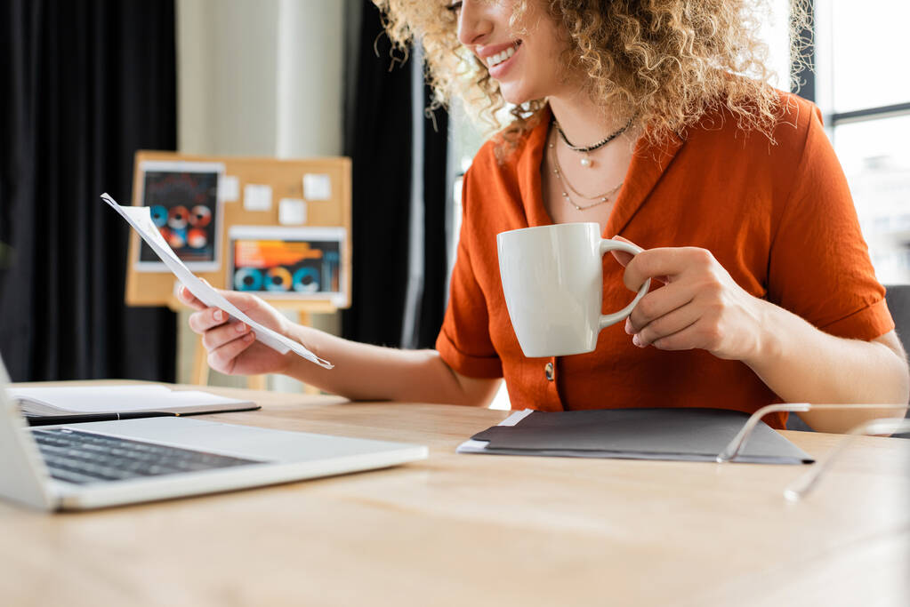 vista recortada de alegre mujer de negocios con pelo rizado sosteniendo taza de té cerca de la computadora portátil  - Foto, imagen