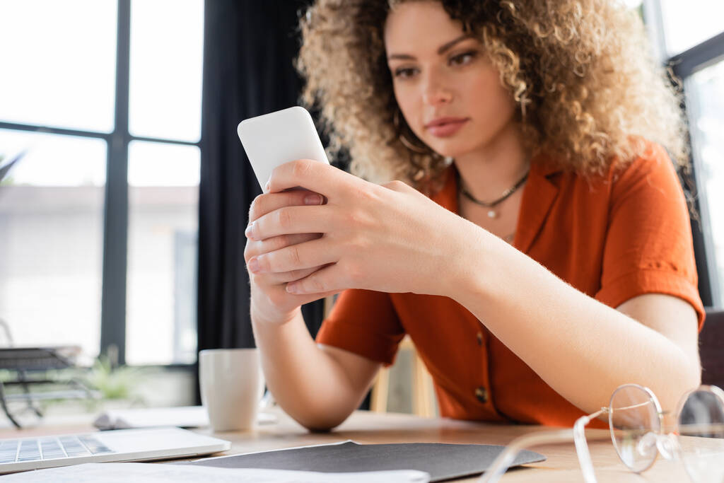 Lockige Geschäftsfrau nutzt Smartphone neben Laptop neben Brille auf Schreibtisch  - Foto, Bild