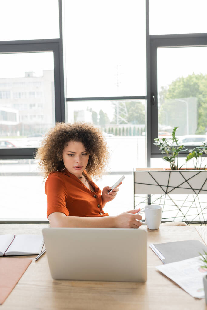 kręcone businesswoman gospodarstwa smartfon i filiżankę herbaty podczas patrząc na laptopa w biurze  - Zdjęcie, obraz