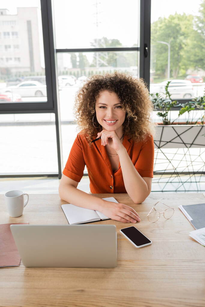 joyful businesswoman with curly hair looking at camera near gadgets and cup of coffee on desk  - Photo, Image
