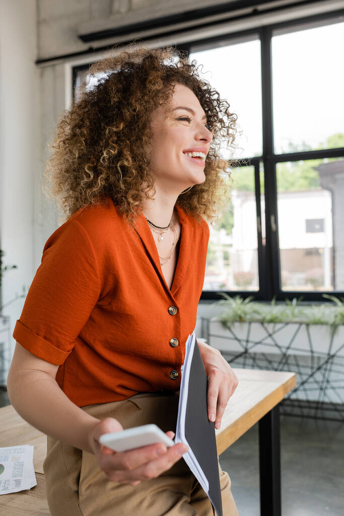allegra donna d'affari con cartella per capelli ricci e smartphone mentre rideva in ufficio  - Foto, immagini
