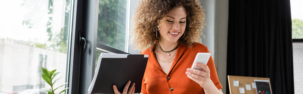 cheerful businesswoman with curly hair holding folder and using smartphone in office, banner - Photo, Image