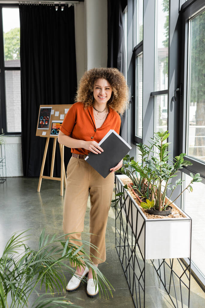 full length of cheerful businesswoman with curly hair holding folder and looking at camera in modern office  - Photo, Image
