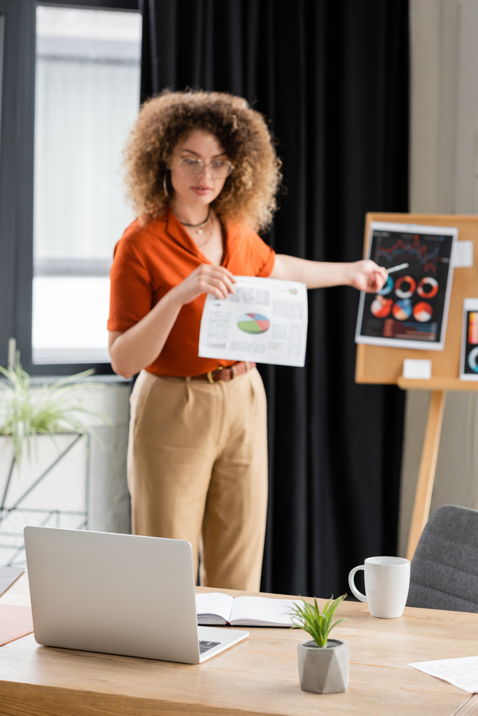 mulher de negócios encaracolada em óculos apontando com caneta para infográficos na placa de escritório durante a chamada de vídeo  - Foto, Imagem