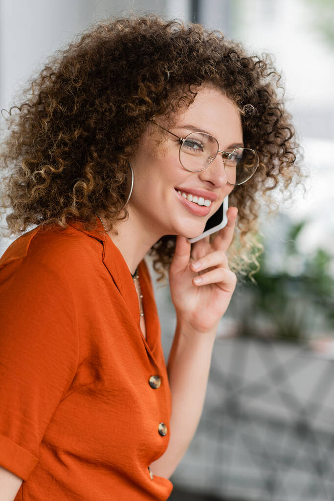 lockige und glückliche Geschäftsfrau mit Brille, die im Büro auf dem Smartphone spricht  - Foto, Bild
