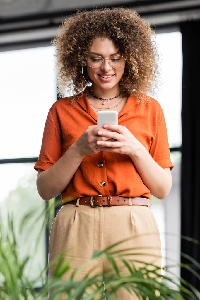 alegre mujer de negocios en gafas de mensajería en el teléfono inteligente, mientras que de pie en la oficina moderna  - Foto, Imagen