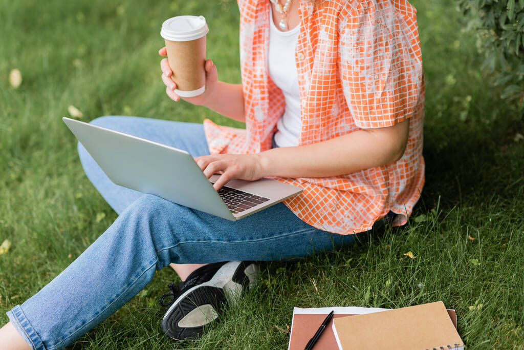 cropped view of young freelancer holding paper cup and using laptop while sitting on grass  - Photo, Image