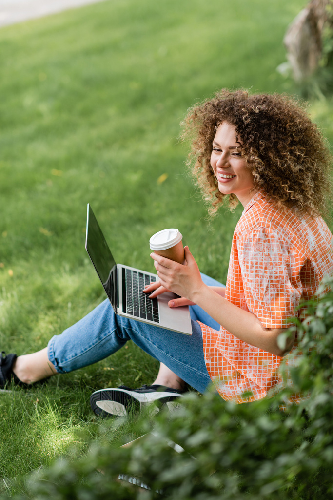 potěšený na volné noze s kudrnaté vlasy drží papírový šálek a pomocí notebooku, zatímco sedí na trávě  - Fotografie, Obrázek