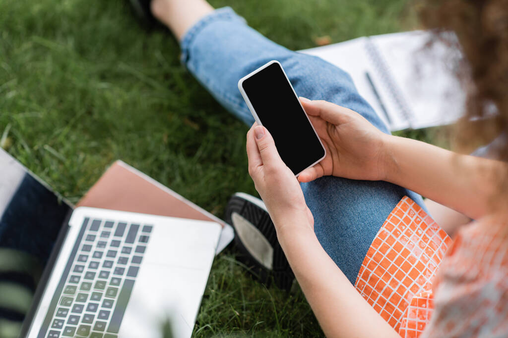 обрезанный вид молодой женщины, держащей смартфон с чистым экраном, сидя рядом с ноутбуком и ноутбуком на траве  - Фото, изображение