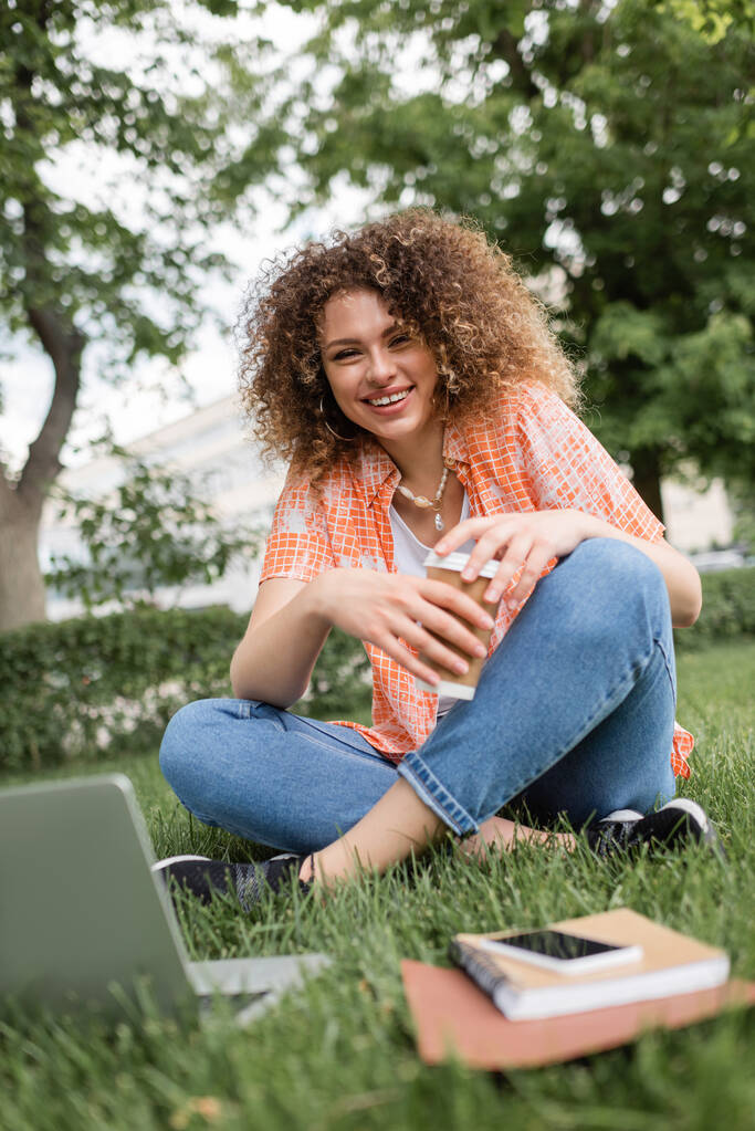 felice donna freelance in possesso di tazza di carta e seduto su erba verde vicino al computer portatile  - Foto, immagini