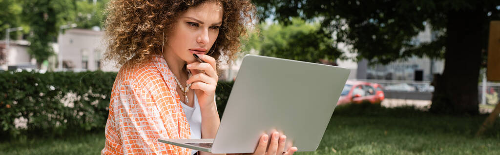 freelancer pensativo con pelo rizado que sostiene el ordenador portátil mientras está sentado en el parque verde, pancarta  - Foto, imagen