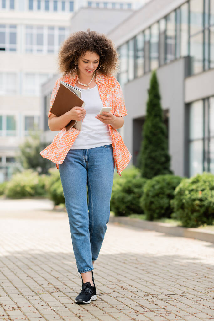 full length of cheerful woman with curly hair using smartphone and holding laptop with folder while walking outside - Photo, Image