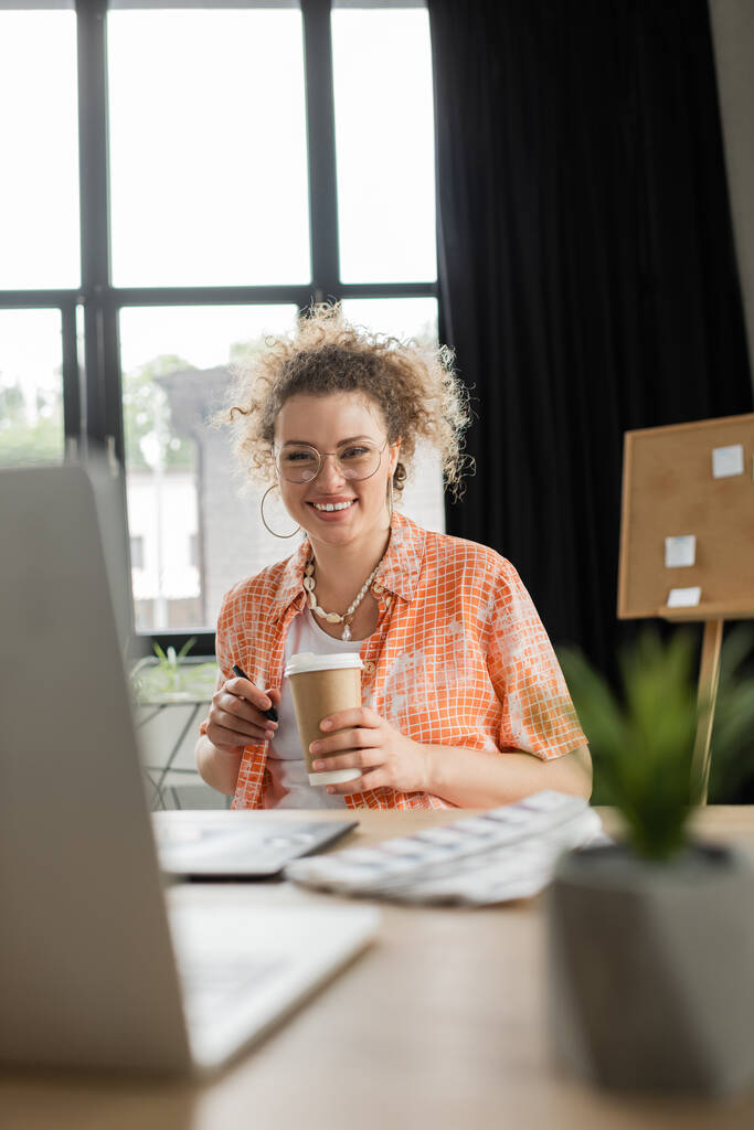 χαρούμενη σχεδιαστής αρχιτεκτονικής σε γυαλιά κρατώντας καφέ για να πάει κοντά gadgets στο γραφείο - Φωτογραφία, εικόνα