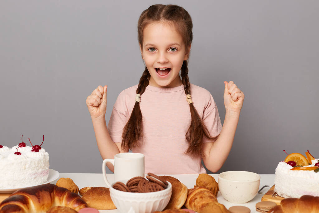 Porträt eines aufgeregten fröhlichen überglücklichen kleinen Mädchens mit Zöpfen, das isoliert am Tisch vor grauem Hintergrund sitzt, die Fäuste geballt und bereit ist, viele Süßigkeiten und hausgemachtes Gebäck zu essen. - Foto, Bild