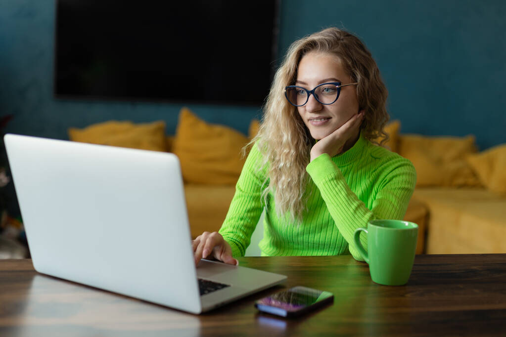 Uma jovem freelancer de óculos olha de surpresa para uma tela de laptop em uma mesa no quarto. Mantém uma xícara verde com uma bebida  - Foto, Imagem