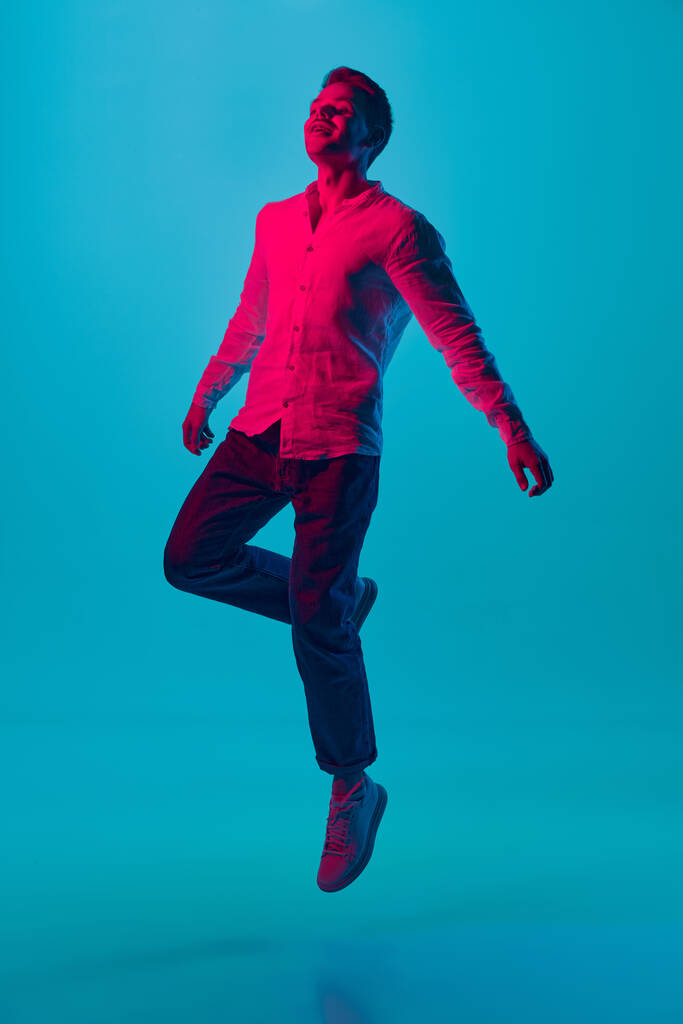 Calm, happy and delightful. Full-length portrait of young positive man posing in jump against blue studio background in pink neon light. Concept of youth, emotions, facial expression, lifestyle. Ad - Photo, Image