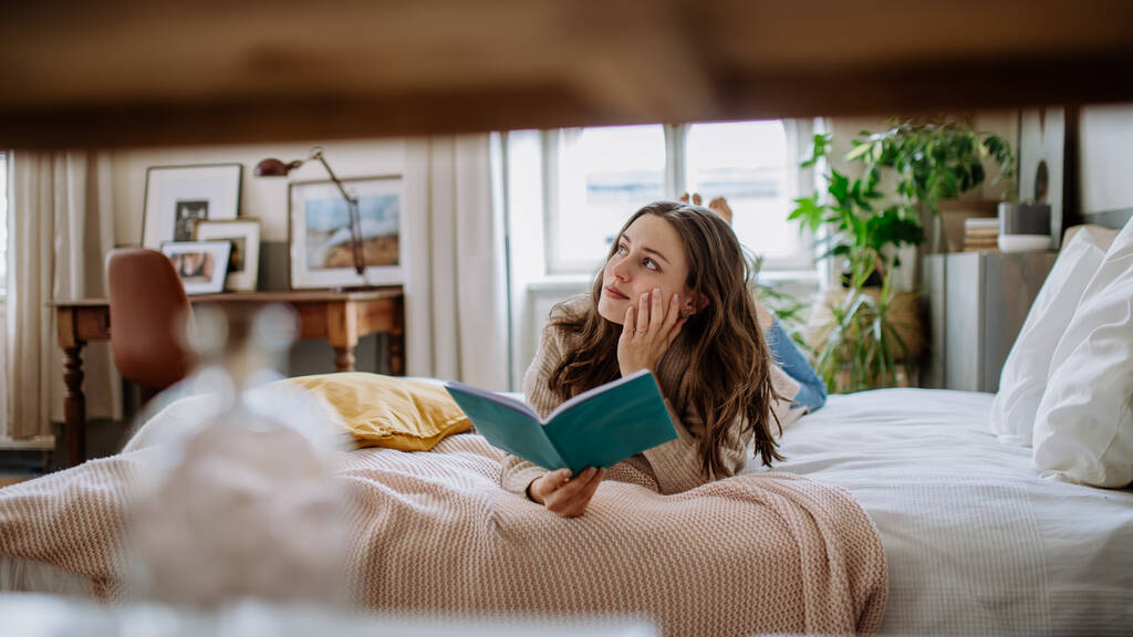 Młoda kobieta ciesząca się wolnym czasem w mieszkaniu, czytając książkę. - Zdjęcie, obraz