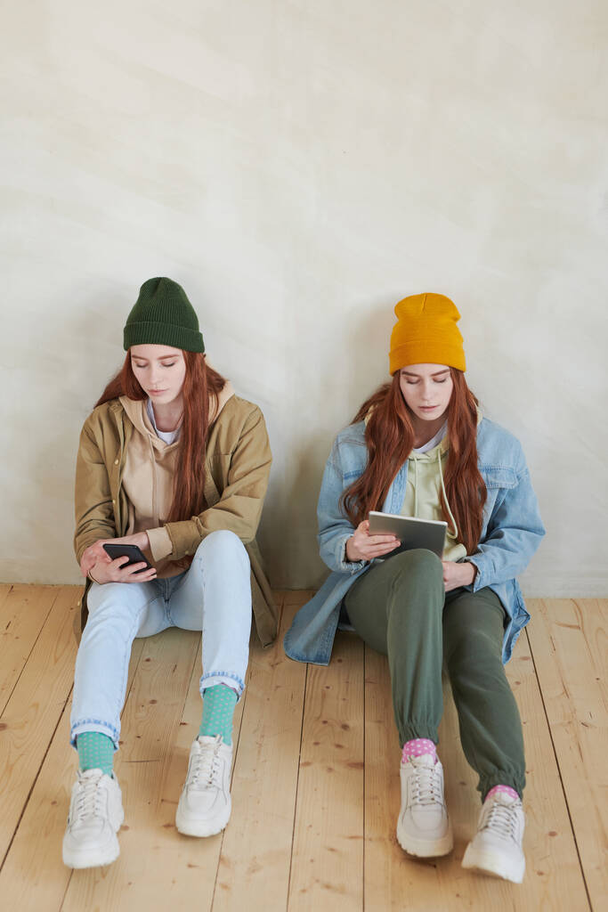 Vista de ángulo alto de hermanas gemelas elegantes que usan ropa casual sentadas en el piso navegando por Internet en sus gadgets - Foto, imagen