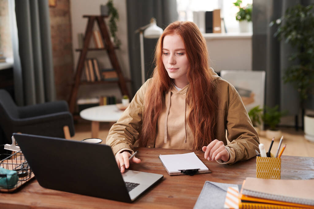 Horizontal medium portrait of female university student with long red hair sitting at desk doing homework using laptop and making notes - Photo, Image