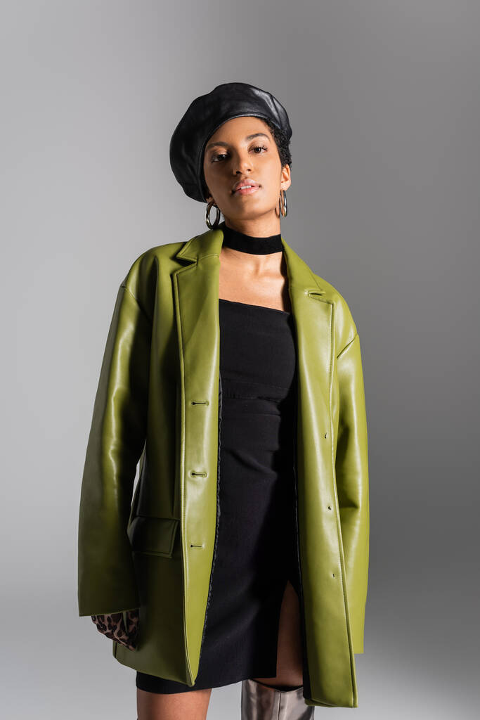 Young african american woman in leather beret and coat looking at camera isolated on grey  - Photo, Image