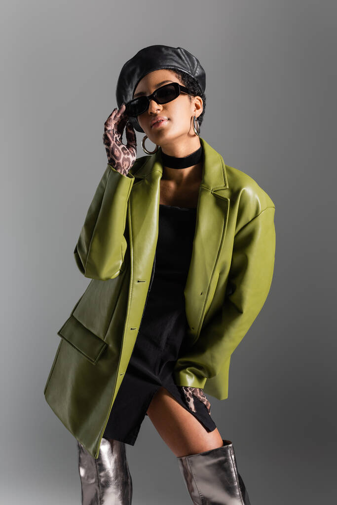 グレーで隔離されたサングラスに触れるレザーコートでファッショナブルなアフリカ系アメリカ人モデル  - 写真・画像