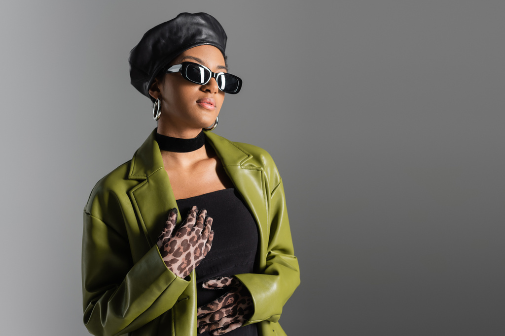 Μοντέρνα Αφροαμερικανή γυναίκα με γυαλιά ηλίου και πράσινο παλτό που στέκεται απομονωμένη στο γκρι  - Φωτογραφία, εικόνα