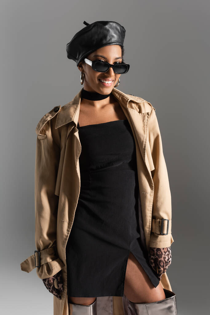 Usmívající se africká americká žena v slunečních brýlích a trenč kabát při pohledu na kameru izolované na šedé  - Fotografie, Obrázek