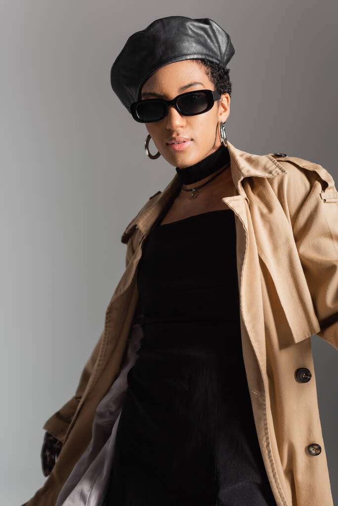 Modelo afroamericano de moda en gafas de sol y gabardina de pie aislado en gris  - Foto, Imagen