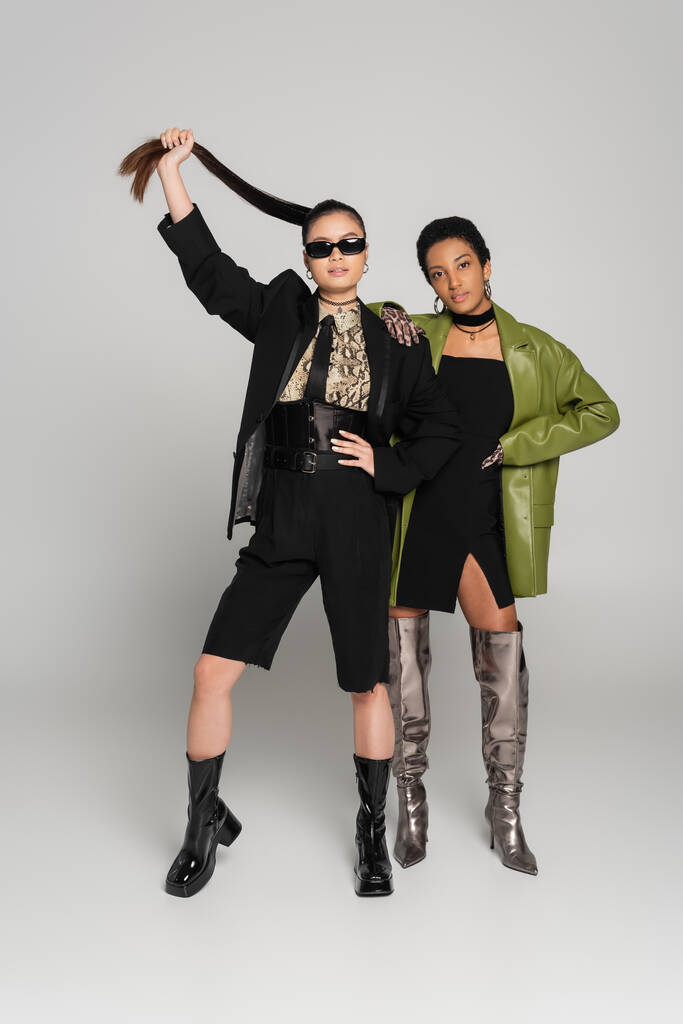 Full length of stylish interracial girlfriends in jackets and boots posing on grey background - Photo, Image