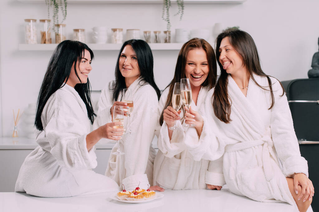 A group of women spend leisure time. Young attractive females in bathrobes drink champagne and chat merrily at home. - Φωτογραφία, εικόνα