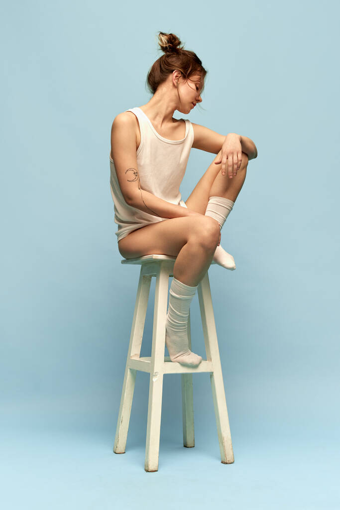 Portret van een mooi, jong, ontspannen meisje met haarknot, in comfortabele huiskleding en sokken poserend op stoel tegen een blauwe studioachtergrond. Lichaams- en huidverzorging, figuur, fitness, gezondheid, wellness-concept - Foto, afbeelding