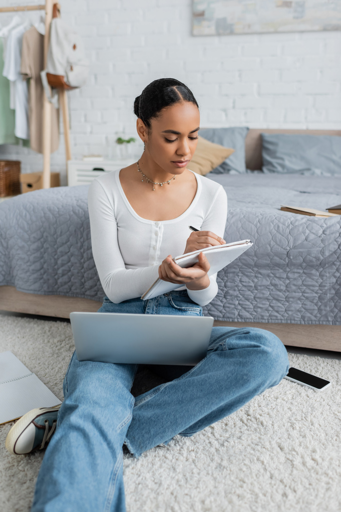 mujer afroamericana bonita en jeans tomando notas mientras ve webinar en el portátil  - Foto, imagen