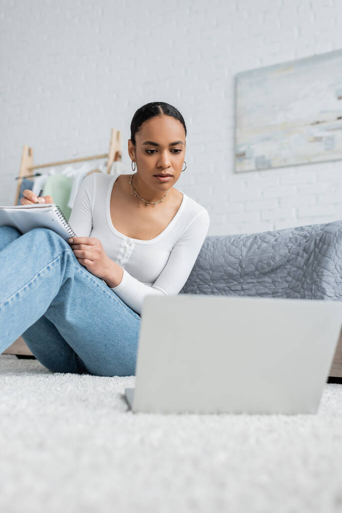 jolie femme afro-américaine en jeans prenant des notes tout en regardant une conférence en ligne sur ordinateur portable  - Photo, image