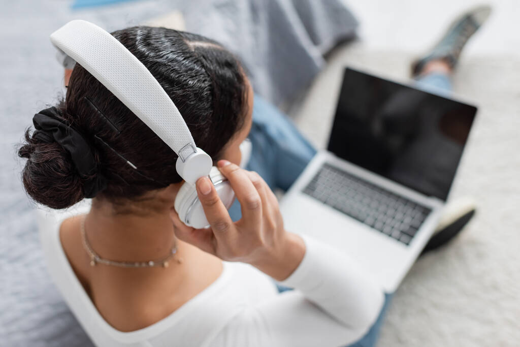 vista superior del estudiante afroamericano que usa auriculares inalámbricos mientras escucha podcast y usa computadora portátil  - Foto, imagen