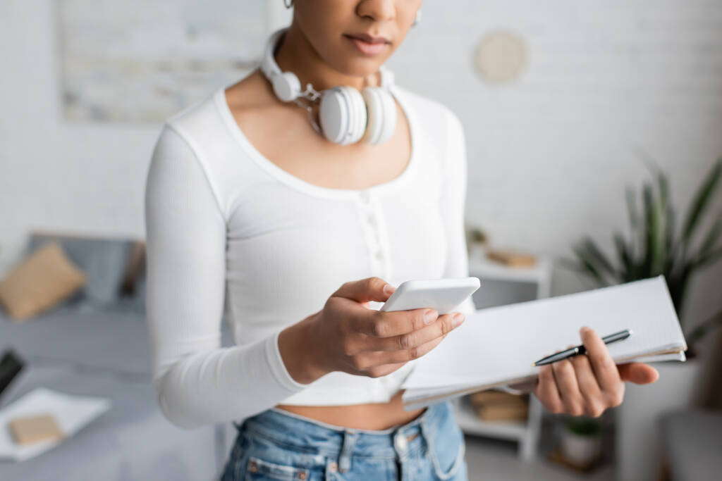 cropped view of african american student with wireless headphones on neck using smartphone and holding notebook  - Photo, Image