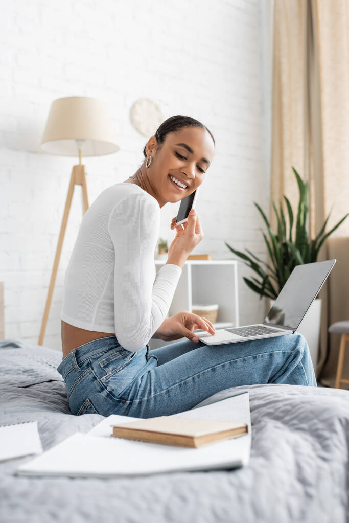 mosolygós afro-amerikai diák beszél okostelefon használata közben laptop és tanulás online otthonról  - Fotó, kép