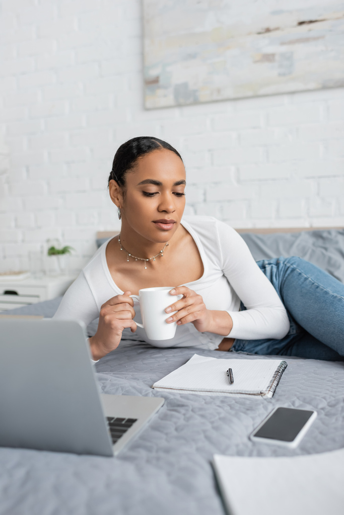 молодая африканская студентка, держит чашку кофе и смотрит на ноутбук рядом с ноутбуком в современной спальне  - Фото, изображение