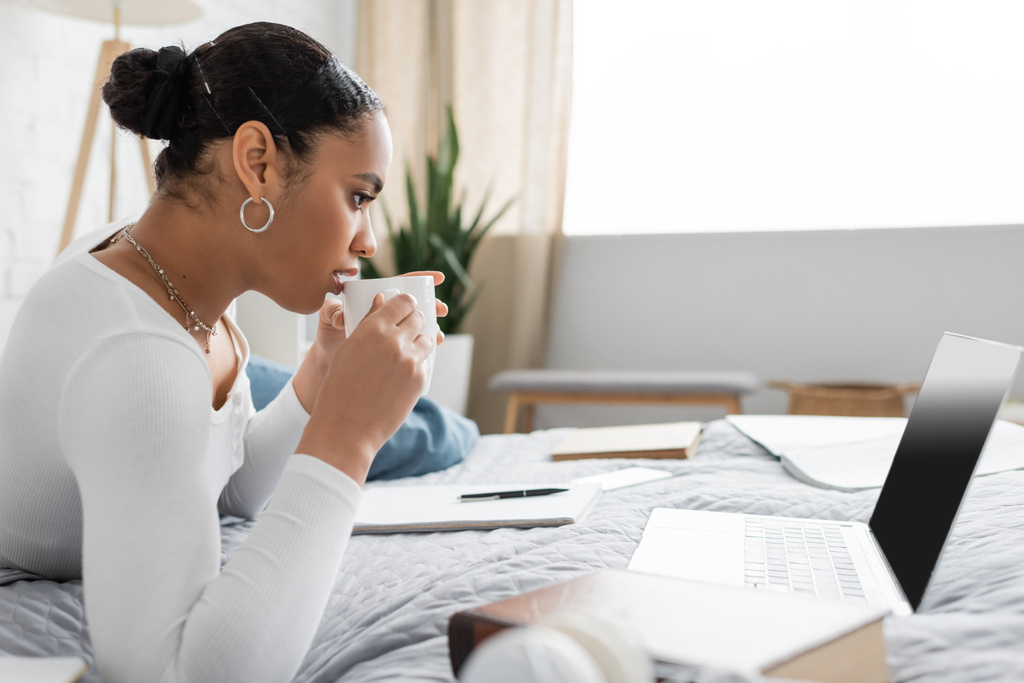Jolie étudiante afro-américaine tenant une tasse de café et regardant un ordinateur portable avec écran blanc  - Photo, image