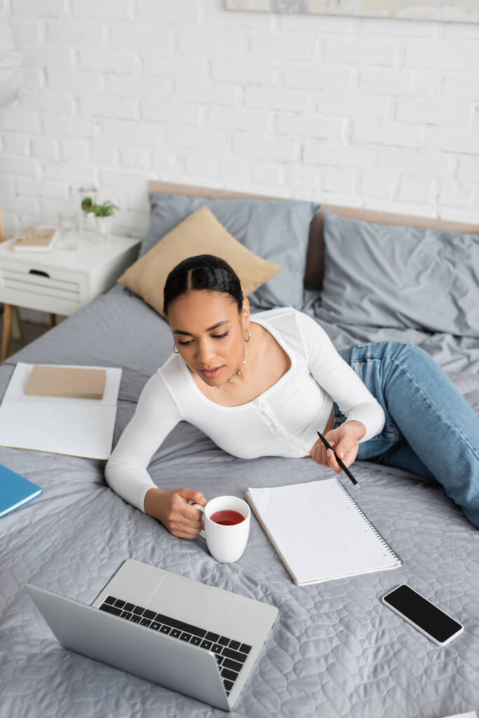 African american student holding cup of tea near laptop and notebook on bed at home  - Photo, Image