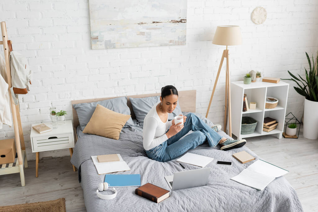 Estudiante afroamericano sosteniendo taza de té cerca de gadgets y libros en la cama en casa  - Foto, Imagen
