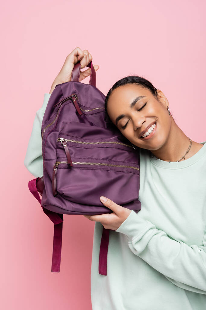 pleased african american student with closed eyes holding purple backpack isolated on pink  - Photo, Image